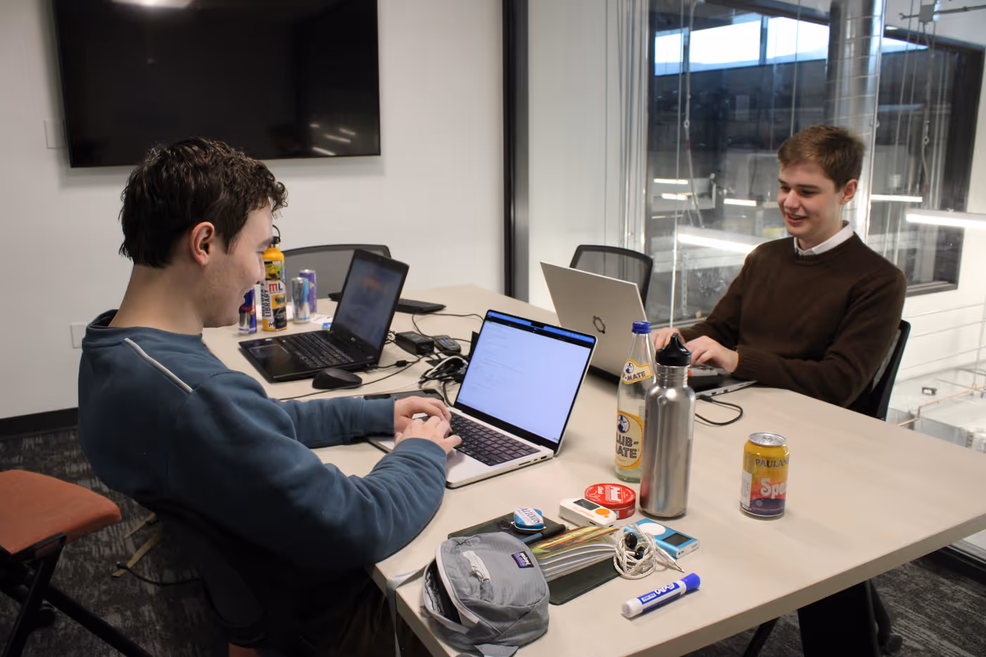 Lukas and Grant write code on their laptops. Beverage cans, electronics, and miscellaneous paraphernalia are strewn across their table.